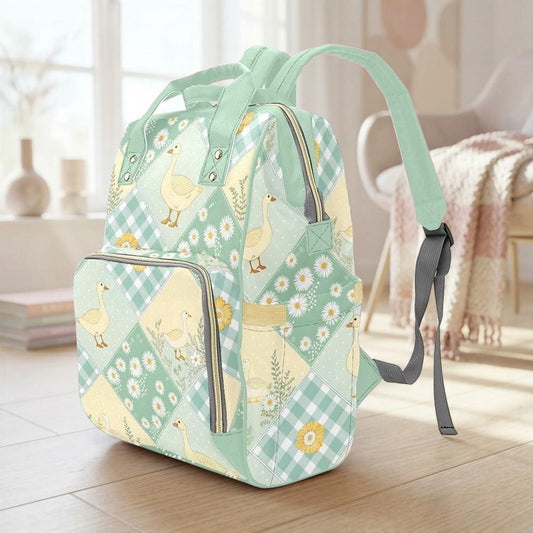 A green and white backpack with a pattern of ducks and flowers is sitting on a wooden floor in a room with a window and a chair.