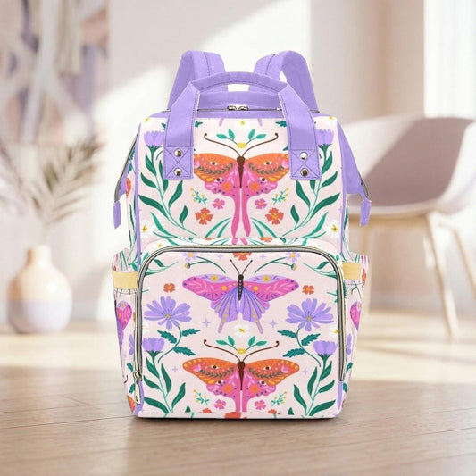 A colorful and vibrant backpack with a floral and butterfly design, featuring purple and pink colors, is displayed on a wooden floor in a room with a white chair and a vase in the background.