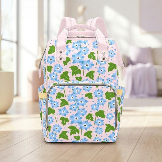 A pink backpack with a floral pattern of blue and green flowers is displayed on a wooden floor in a room with a window.