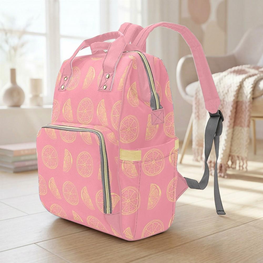 A pink backpack with a pattern of lemon slices is sitting on a wooden floor in a room with a window and a chair.