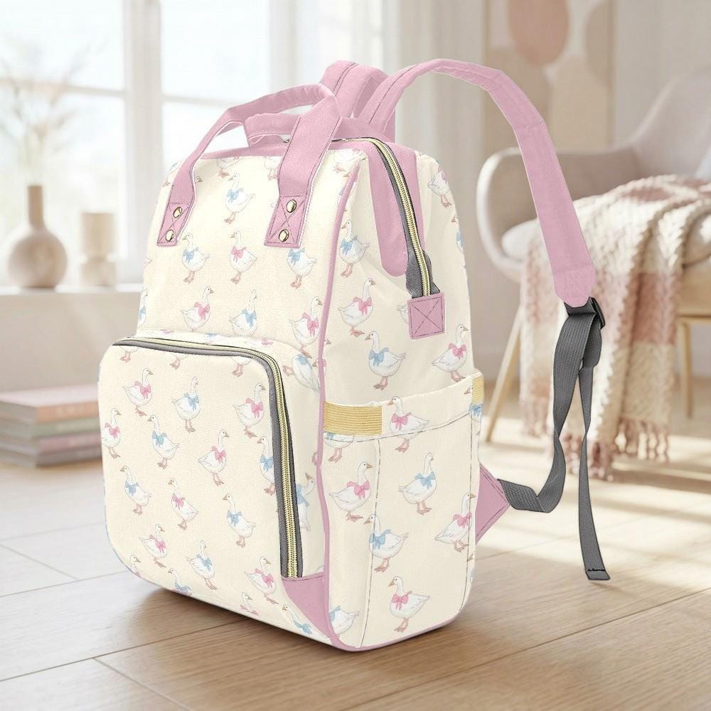 A pink and white patterned backpack with ducks printed on it, sitting on a wooden floor in a room with a window and a chair.