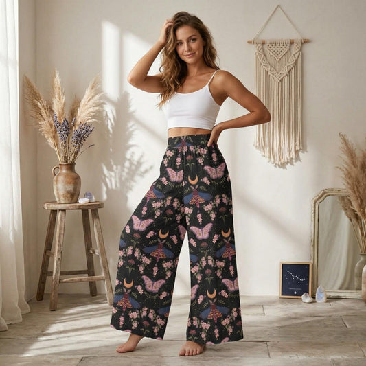 A woman wearing a white crop top and wide-leg pants with a floral print stands in a room with a wooden stool, a vase of flowers, and a macrame wall hanging.