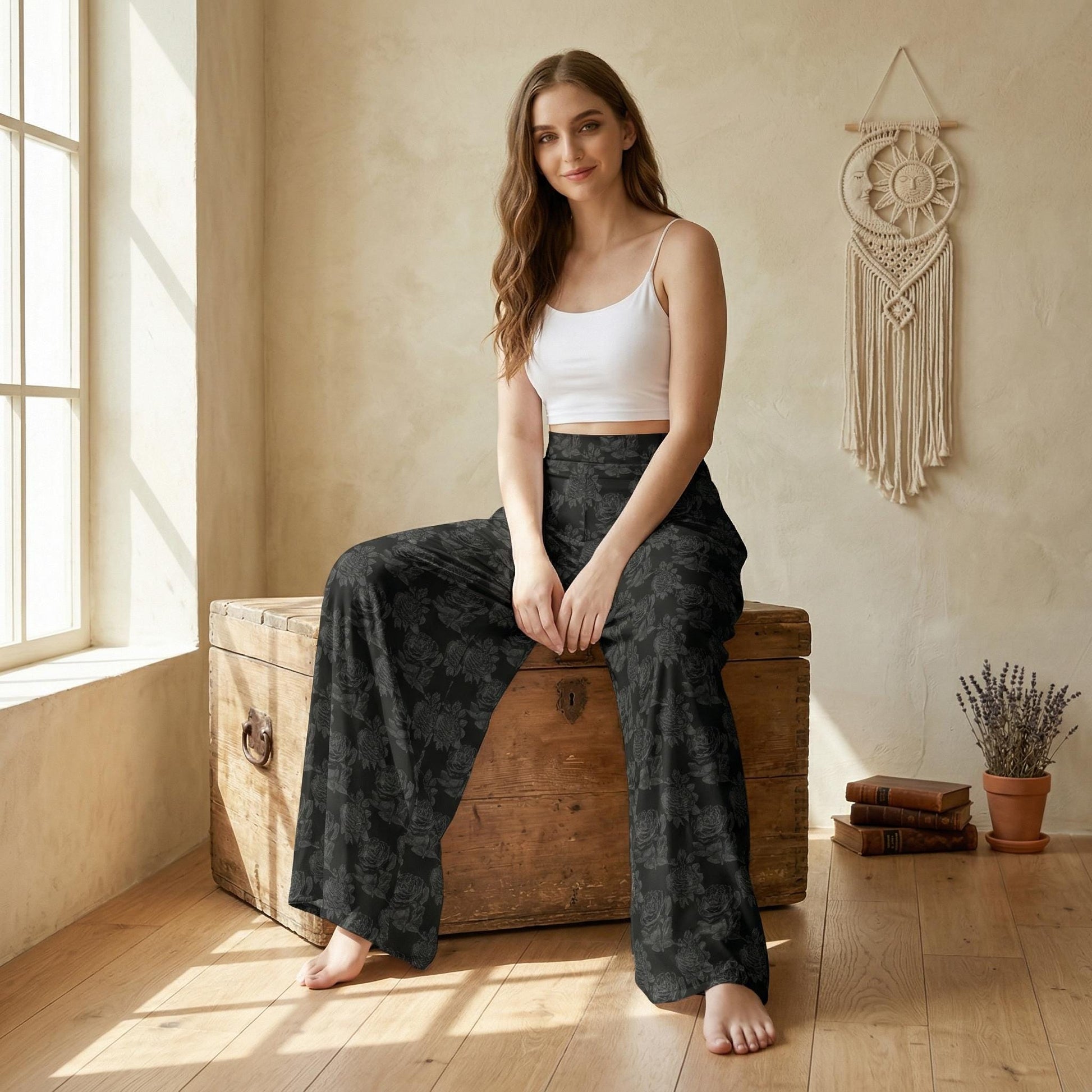 A woman in a white crop top and black floral pants sits on a wooden trunk in a room with a window and a hanging dreamcatcher.