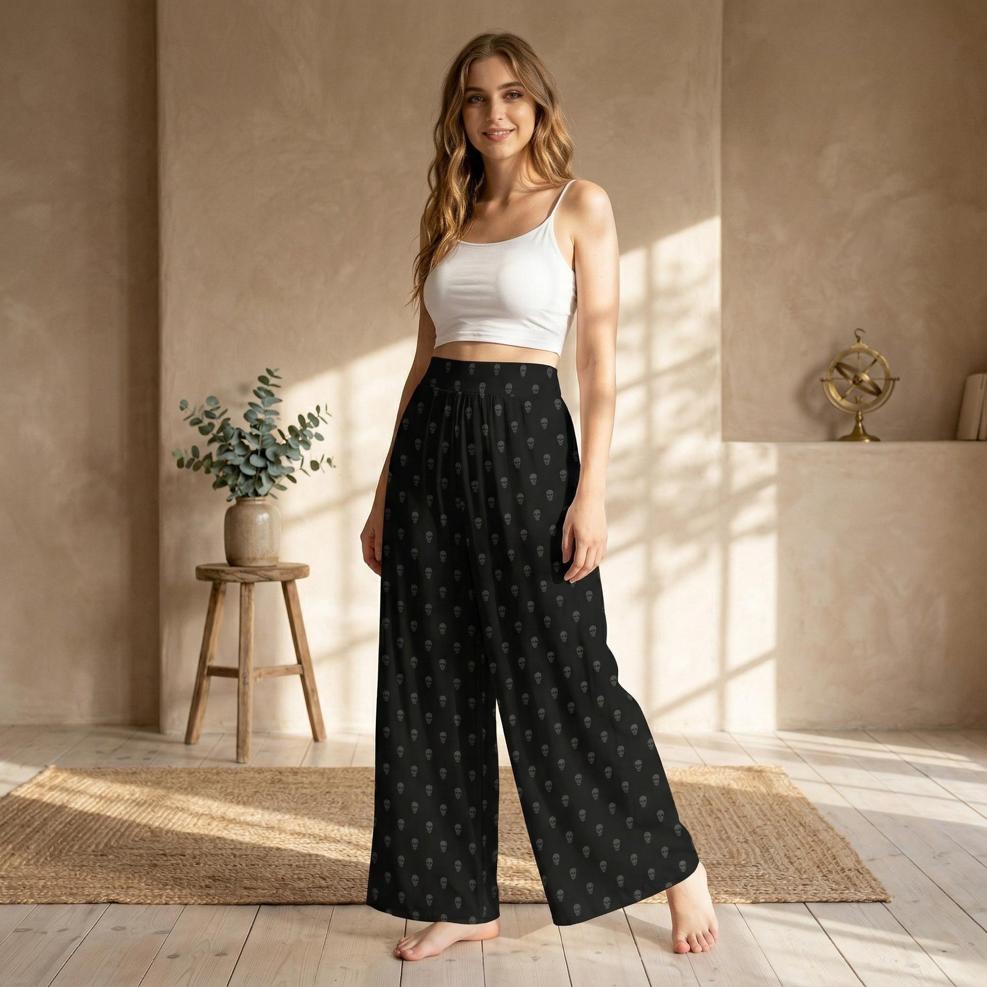 A woman wearing a white crop top and black patterned wide-leg pants stands in a room with a wooden stool, a vase of greenery, and a gold-colored object on a shelf.