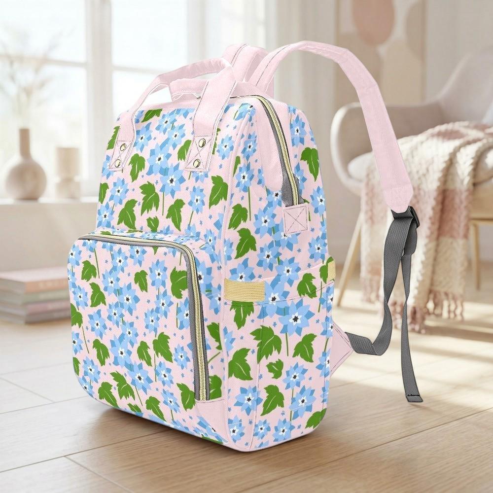 A pink and green floral backpack is sitting on a wooden floor in a room with a window and a chair visible in the background.