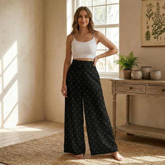 A woman wearing a white crop top and black patterned wide-leg pants stands in a room with a wooden table and potted plants.