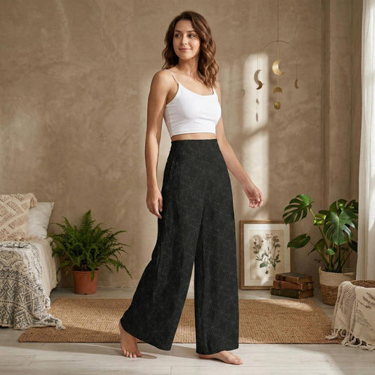 A woman wearing a white crop top and black patterned wide-leg pants stands in a room with a beige wall, potted plants, and a framed picture.