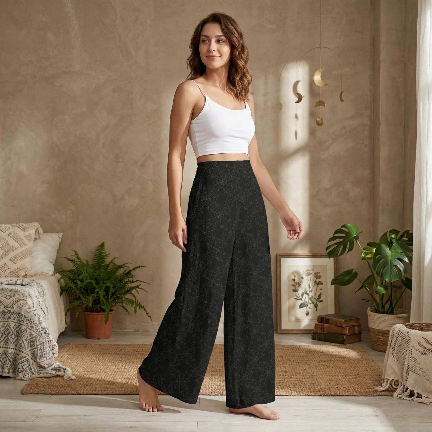 A woman wearing a white crop top and black patterned wide-leg pants stands in a room with a beige wall, potted plants, and a hanging crescent moon decoration.