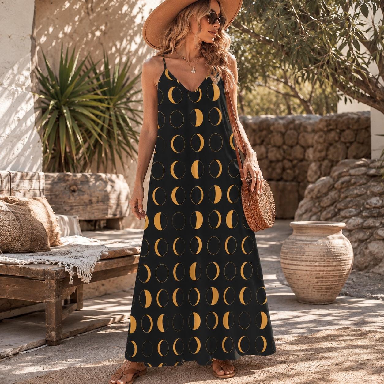 Woman wearing a long black spaghetti strap maxi dress printed all over with large mustard yellow moon phases, holding a round rattan bag on a sunlit stone patio.