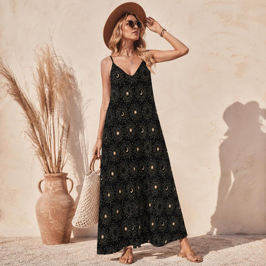 Woman wearing a long black spaghetti strap maxi dress printed all over with gold and cream moon phase orbs and tiny stars, holding a woven bag beside a terracotta pot.