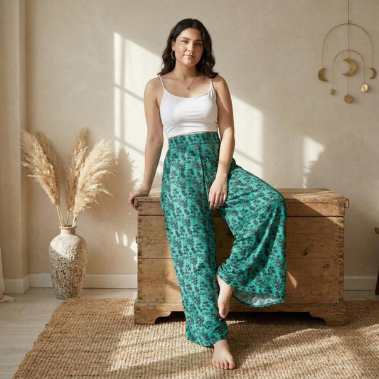 A woman in a white tank top and green floral pants is posing on a wooden chest in a room with a beige wall, a potted plant, and a hanging decoration.