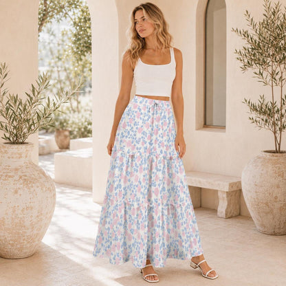 A blonde model stands wearing a white tank top and a long tiered floral maxi skirt with pink roses and blue leaves.